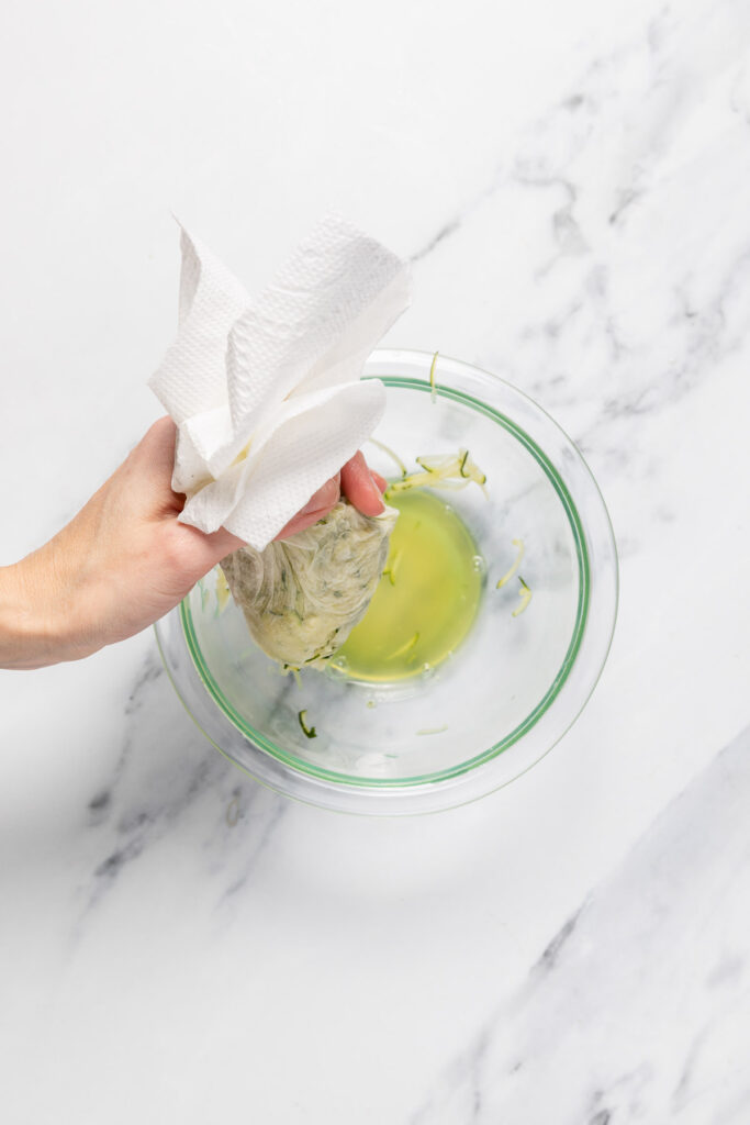 grated zucchini moisture being squeeze onto a bowl with a couple paper towels