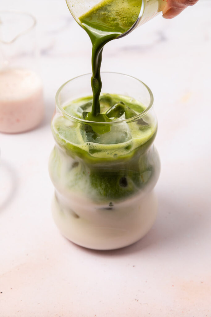 matcha mixture being poured in a tall glass with oat milk